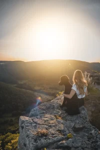 Eine blonde Frau sitzt mit einem schwarzen Pudel bei einem Sonnenuntergang auf einem Felsen.